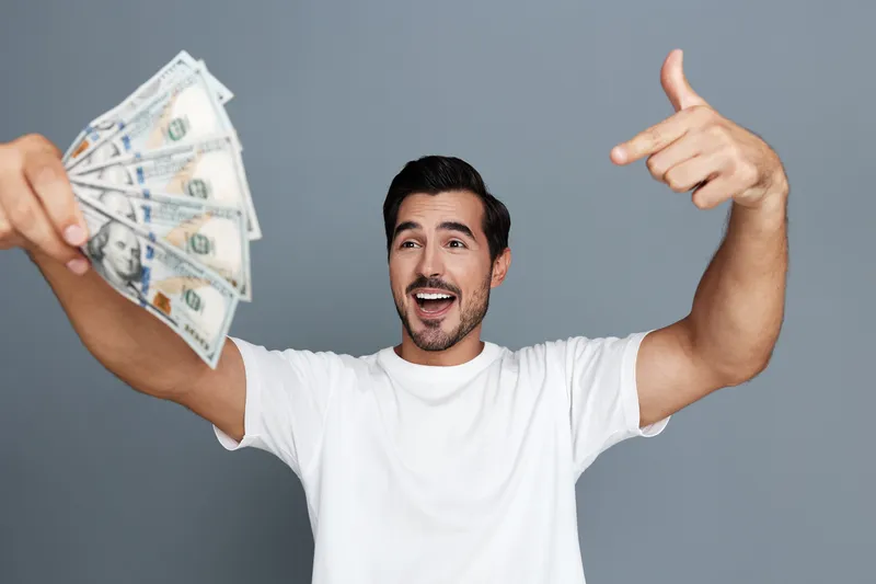 Smiling man holding fan of dollar bills, excited expression with open mouth, dressed in casual white t-shirt, standing against plain gray background. Lifestyle concept.
