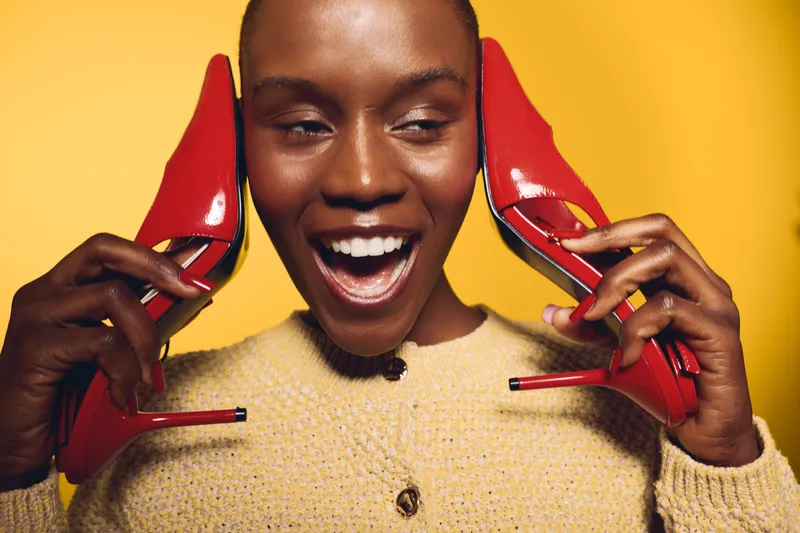 Happy African woman with dark skin holds red high heels near ears smiling brightly. Yellow background and cozy beige sweater create cheerful and stylish fashion concept image.