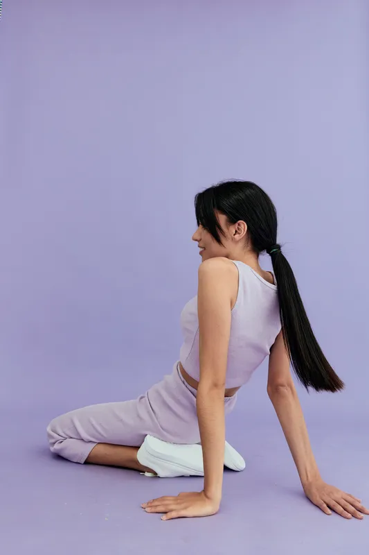 Young woman sitting on the floor in casual lavender outfit and white sneakers against a pastel purple background with relaxed and thoughtful expression.
