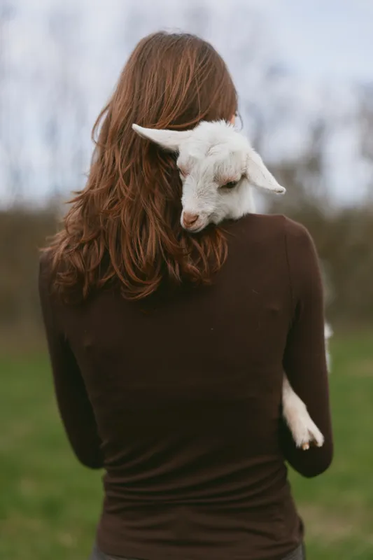 A woman carries a gentle kid on her back in an open field, illustrating care, companionship, and a calm outdoor lifestyle, showcasing the bond between human and animal in a pastoral setting.