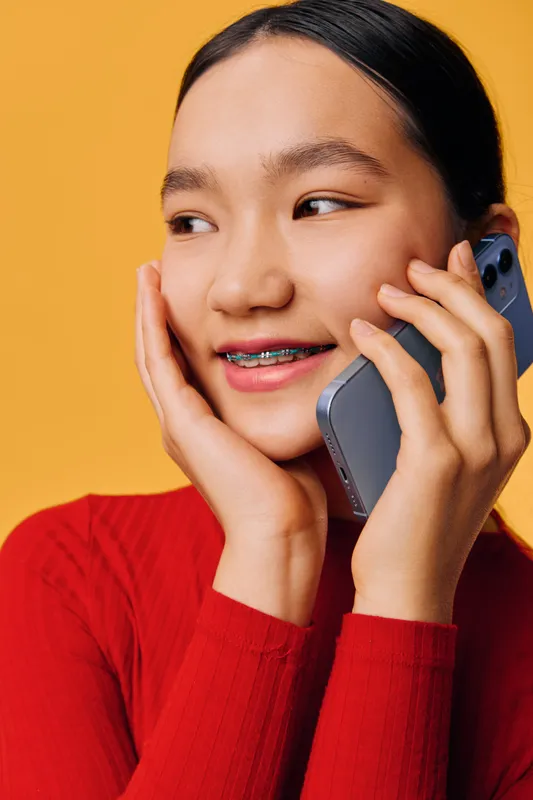 Young woman with braces wearing red sweater talking on smartphone and smiling, closeup portrait with hand on face against yellow background, communication and technology concept
