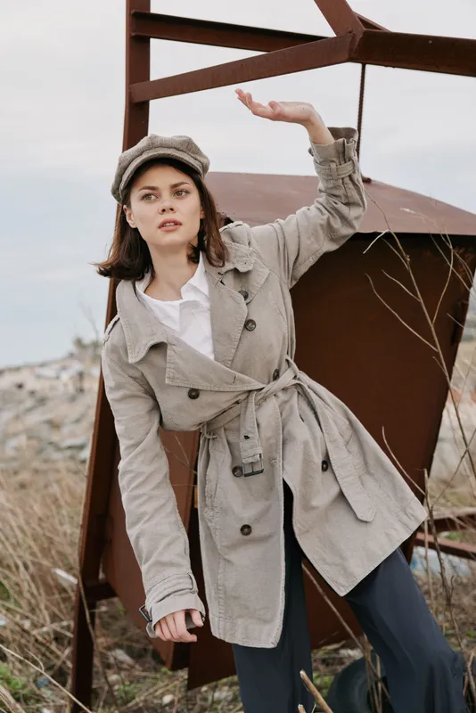 Young woman in a plaid trench coat and matching hat poses outdoors near rusty metal structure. Autumn fashion concept with casual style and natural background in soft daylight.