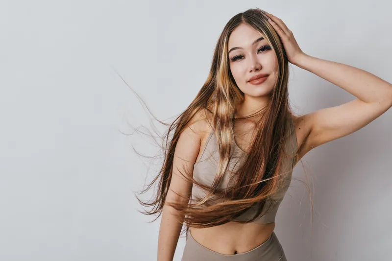 Young woman with long hair posing and smiling in studio, wearing beige crop top, hand on head, natural makeup with closed eyes and casual look on gray background.