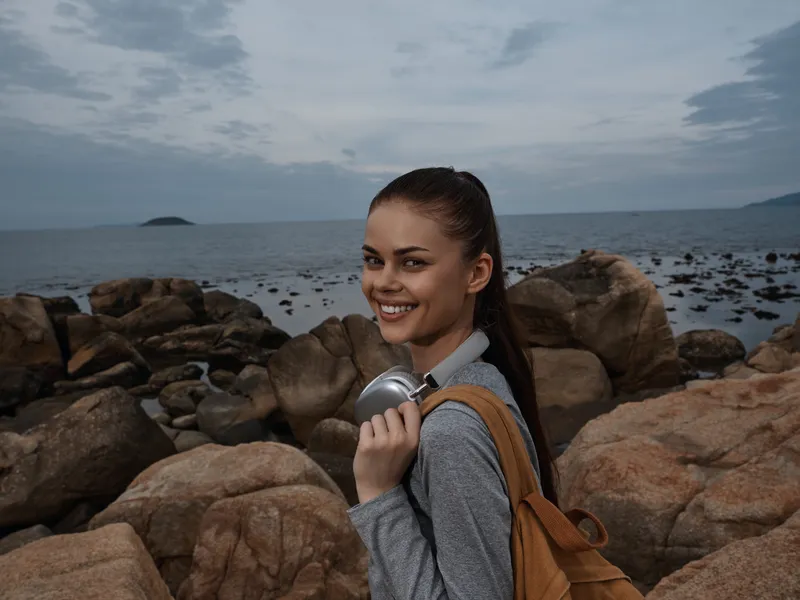 Smiling young woman in casual attire holding a travel mug by the beach, showcasing joy and adventure against a scenic coastal backdrop