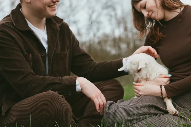 Happy couple outdoors with a gentle goat kid, bonding moment, casual lifestyle, warm smiles, peaceful park scene, pets and nature in harmony.