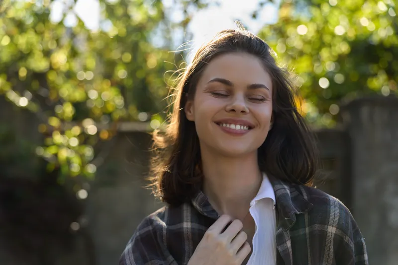 Smiling woman with long hair wearing a plaid shirt in bright outdoor light. Close-up on a natural background, conveying wellness, happiness, and a carefree lifestyle