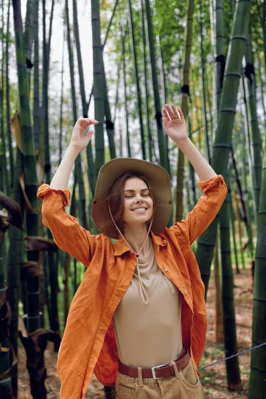 woman bamboo hat smile nature forest travel portrait with joyful relaxed pose in a bamboo grove, wearing orange shirt and wide brim hat, sunlight filtering through green trees for outdoor lifestyle