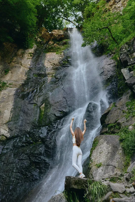 woman practicing yoga near waterfall in lush forest with rocks and green trees, wearing white leggings and top, peaceful outdoor exercise, healthy lifestyle