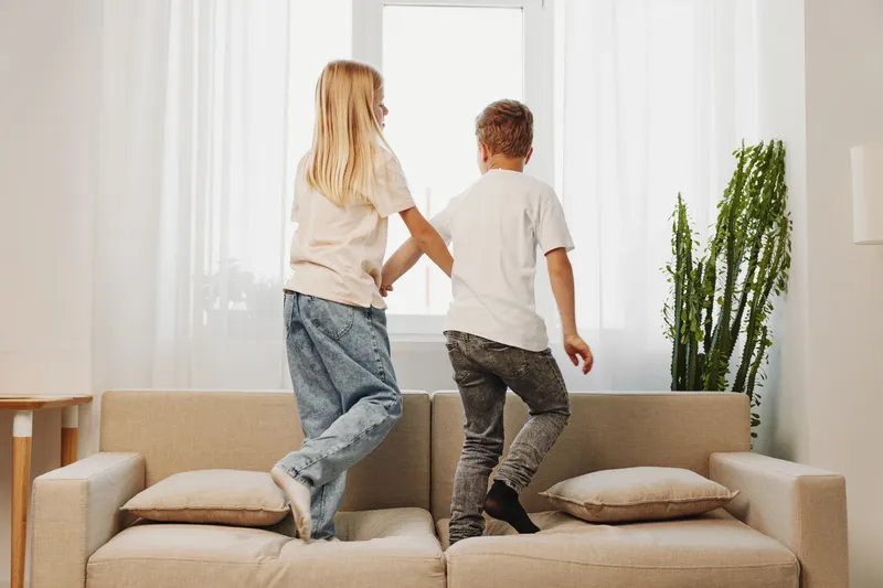 Two children jumping on beige sofa in bright living room with natural light. Siblings have fun together, casual clothes, joyful activity in cozy home environment with plants and cushions.