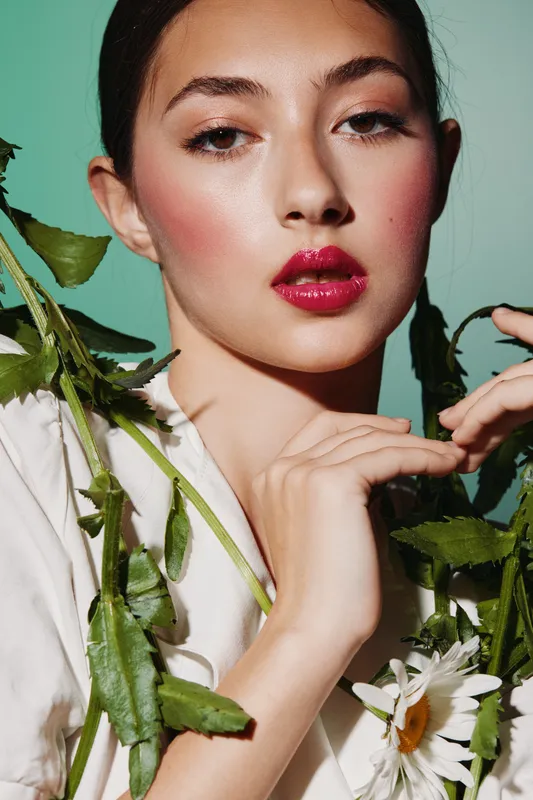 Young woman with creative pink makeup and bold red lips posing with green leaves and white daisies against a pastel background. Concept for beauty, fashion, and artistic style.
