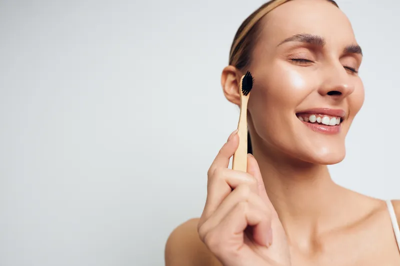 Young woman with clear skin gently cleanses face with a soft facial brush and smiles with closed eyes enjoying skincare routine isolated on white background.