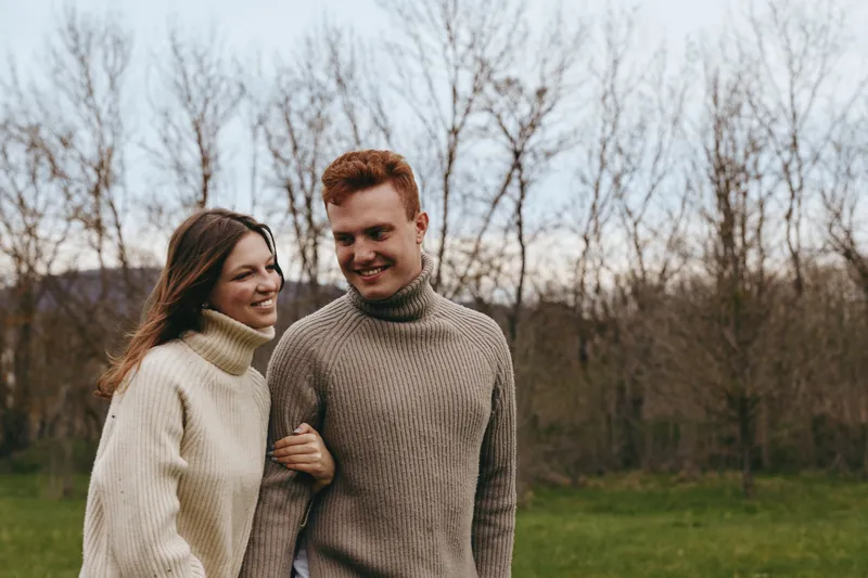 Young couple in cozy sweaters walks arm in arm through a bare tree forest during autumn, sharing a warm and joyful moment outdoors in casual knitwear.