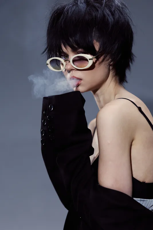Stylish woman with short black hair wearing white sunglasses and black clothing smoking against gray background