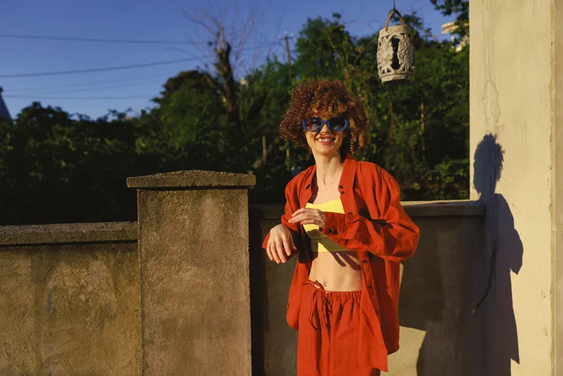 Cheerful woman outdoors, wearing orange lounge set, standing on a sunny terrace, relaxed pose, warm light, casual fashion, carefree mood, blue sky, natural backdrop, playful moment