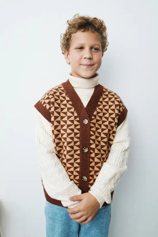A young boy stands against a light wall, wearing a patterned knit vest over a cream turtleneck, with a calm smile and relaxed pose.