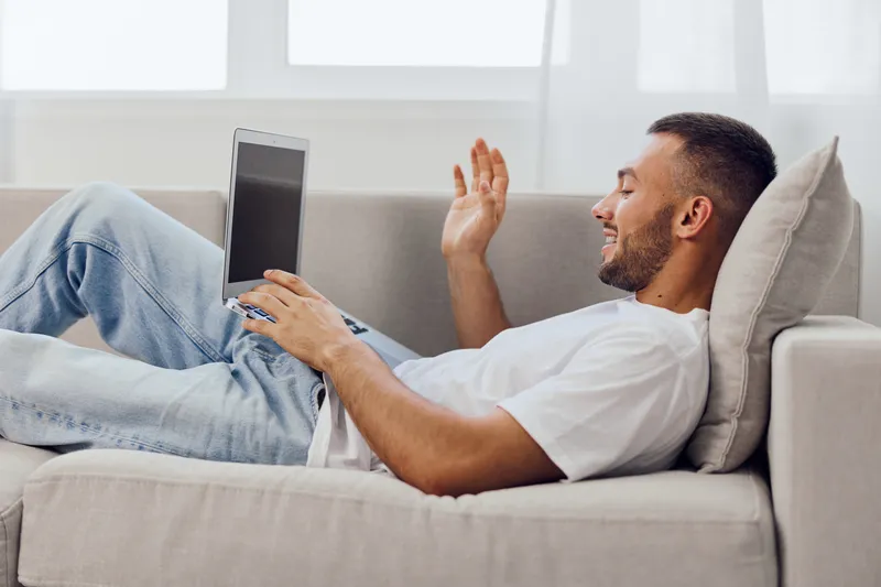 A relaxed man engaging in a video call on a laptop while lying on the couch, representing modern work-life blur and comfort in solo living.