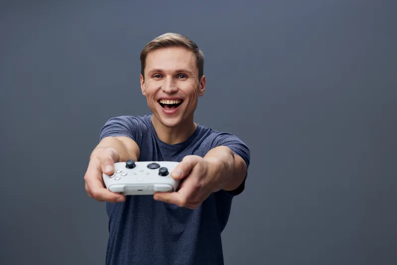 Smiling young man with short brown hair wearing casual blue t-shirt holding game controller and playing video games, isolated on plain gray background. People lifestyle concept, entertainment, fun, te