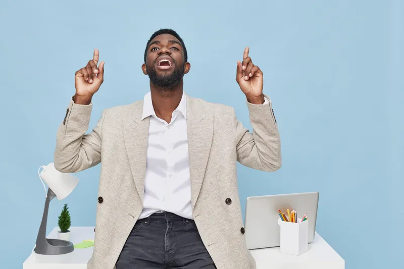 Confident smiling man of African descent in professional attire enjoying success, pointing upwards with both hands, sitting at modern office desk with desktop lamp and supplies on blue background. Bus