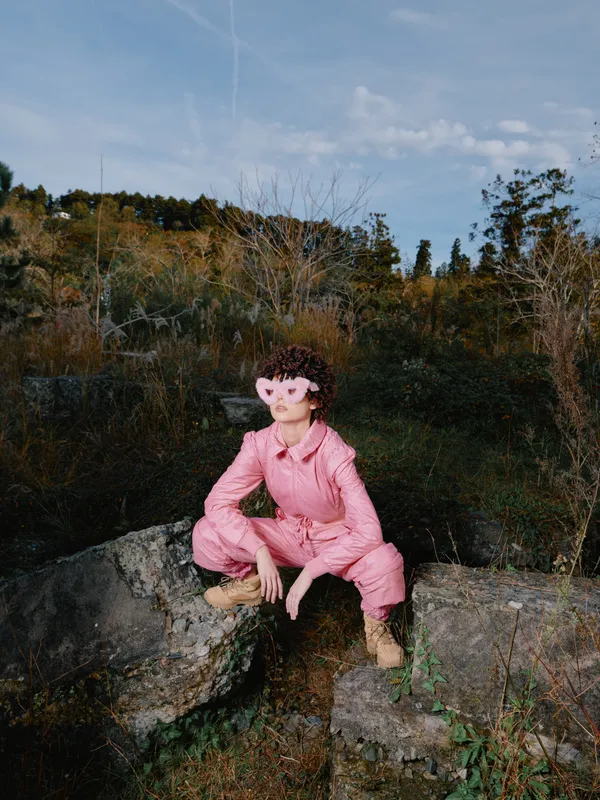 Person outdoors pink mask jumpsuit rock nature landscape pose daytime, a playful portrait in a rugged setting with trees and blue sky, emphasizing bold color and adventurous style.
