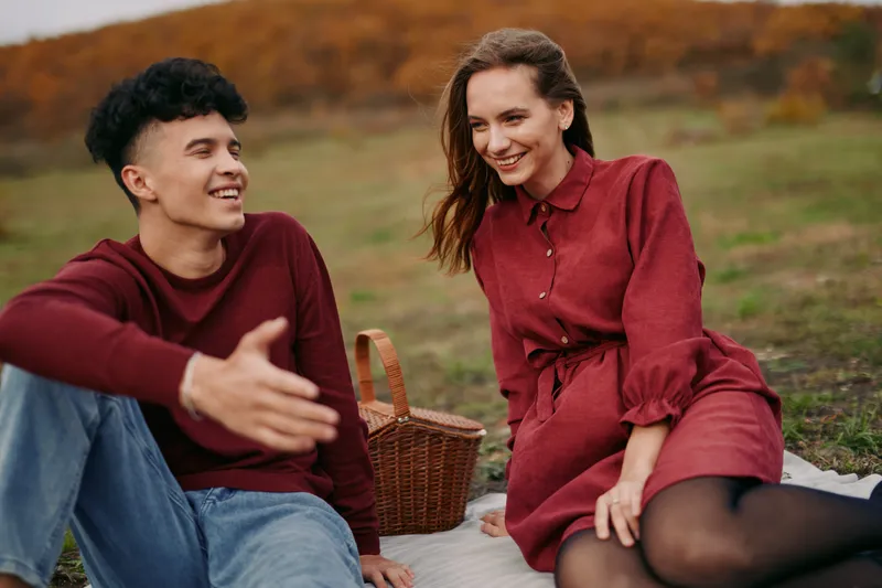 Young couple sharing a genuine outdoor picnic moment, laughing and connecting on a blanket in autumn grass, photographed in a believable, natural candid style for lifestyle use.