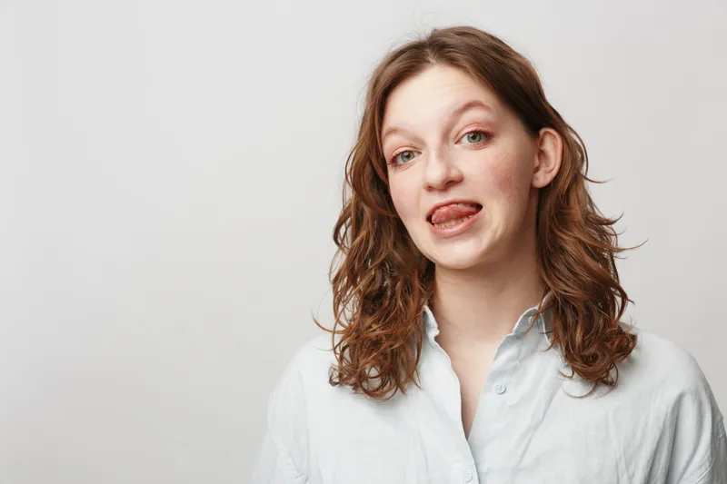 Young woman with curly hair wearing white shirt makes funny face sticking out tongue. Studio portrait on light background. Concept of humor, personality, and casual expression.