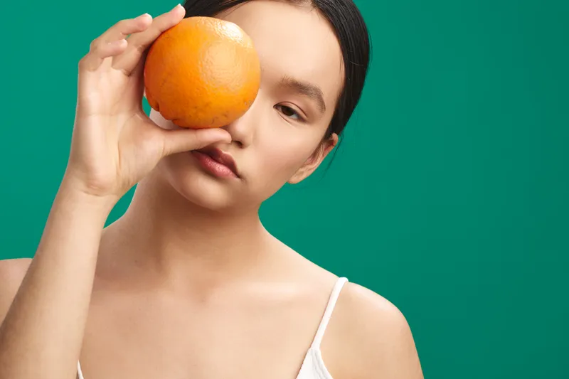 Asian woman holds an orange to her eye in a close up portrait, with a vivid green background, calm expression, natural makeup, and a clean contemporary aesthetic conveying a fresh, playful mood.