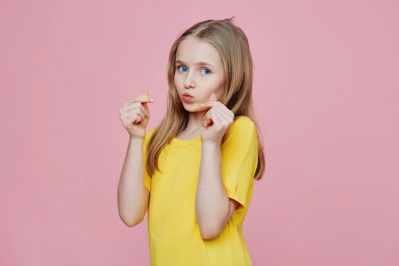 Cheerful girl in a yellow shirt playfully making a pouty face against a pink background, showcasing a fun and youthful energy perfect for children s themes