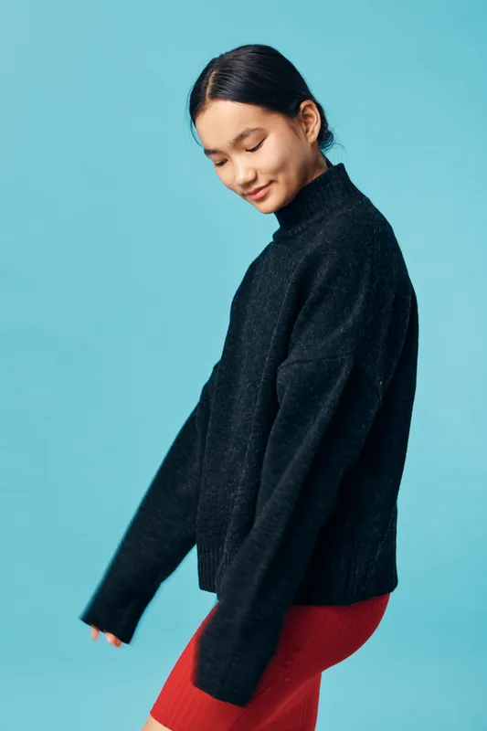 Woman wearing black turtleneck sweater and red skirt posing with eyes closed against blue background, casual fashion portrait indoors with relaxed and peaceful expression.