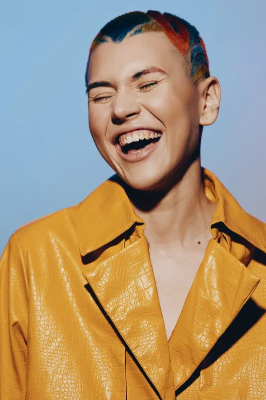 laughing woman with colorful hair and yellow jacket expressing joy and happiness, isolated on blue background with vibrant and playful style.