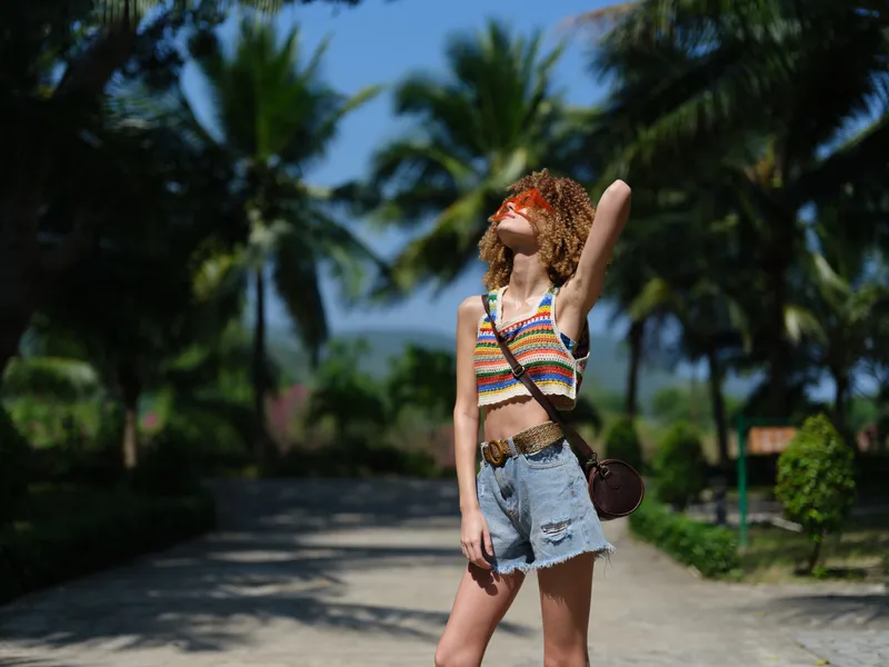 young woman with curly hair wearing colorful striped top and denim shorts standing outdoors, enjoying sunny weather with palm trees and clear sky in background