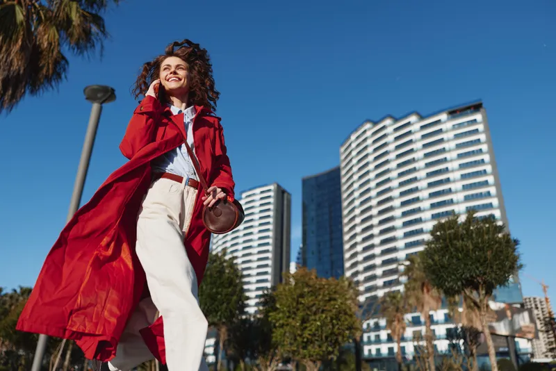 Smiling woman in a red coat walking outdoors beside tall buildings under a clear blue sky, embodying urban fashion and confidence, bright natural lighting, candid street moment.