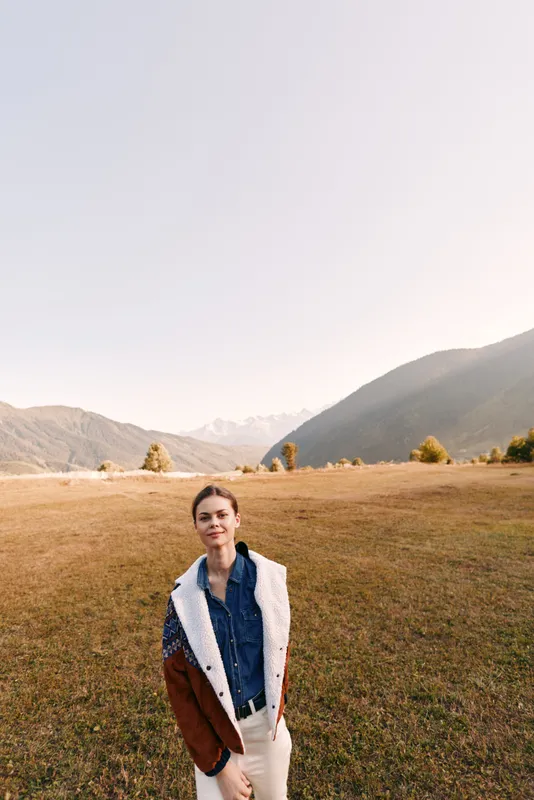 Woman portrait outdoors in a mountain meadow beneath distant mountains, autumn fashion with cozy jacket and relaxed smile. Young female model standing in wide landscape with soft natural light.