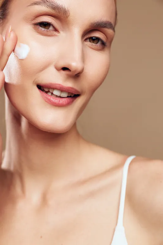 Young woman applies cream to her cheek in a gentle skincare moment, smiling softly as a light glow highlights clear skin in a calm intimate setting