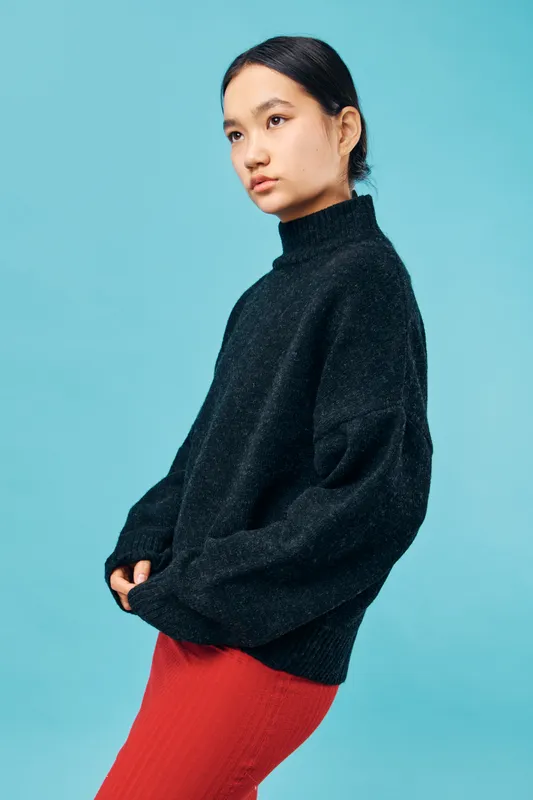 Young woman wearing black oversized sweater and red pants standing against blue background, looking sideways with natural expression and loose hair tied back.