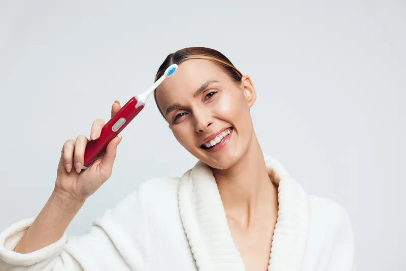 Smiling woman in white bathrobe holds red electric toothbrush near head showing dental hygiene routine isolated on light background with positive facial expression and clean teeth.