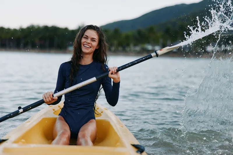 Smiling woman kayaking on calm water, wearing navy blue swimwear, splashing water with paddle, enjoying outdoor adventure and nature.