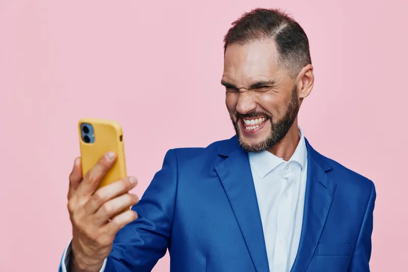 Professional man in a blue suit shares a confident smile while taking a selfie, set against a solid pink background—a dynamic moment of success, focus, and positive energy.
