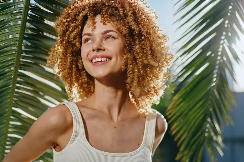 smiling woman with curly hair enjoys sunny warm day outdoors near tropical palm leaves, wearing white tank top and expressing happiness and lightheartedness