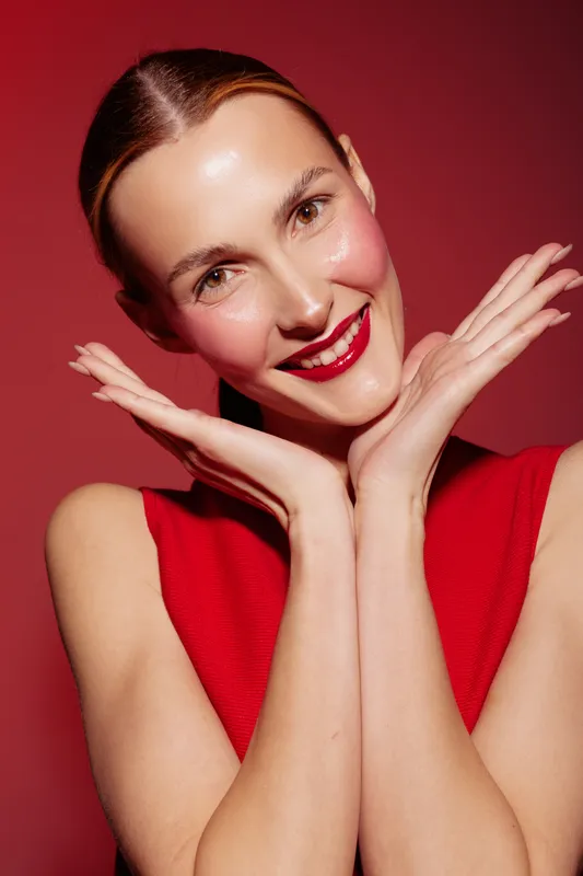 Smiling woman with red lipstick and red sleeveless top poses with hands framing face on red background. Concept of beauty, makeup, fashion, and joyful expression in studio lighting.