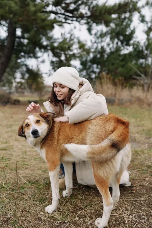 Woman in a warm coat kneels in a grassy field, stroking a friendly dog as soft light filters through trees, capturing a tender moment of companionship and outdoor exploration