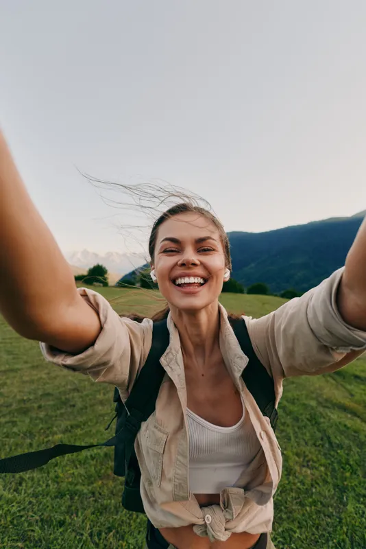 Joyful young woman with a backpack spreads her arms in a wide smile on a grassy field with distant mountains under a clear sky, celebrating an outdoor moment.