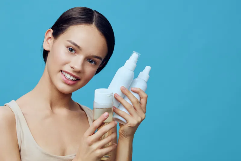 Young girl holding skincare products with a smile, representing healthy skin and beauty care in a bright blue background