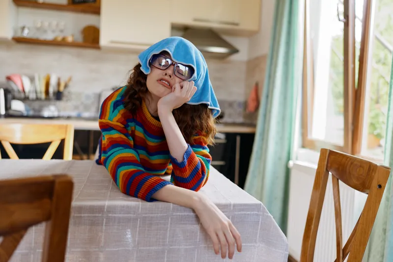 Young woman with curly hair sits at a home dining table, resting her chin on her hand while a blue towel covers her head and oversized sunglasses hide her eyes in a playful, dreamy moment