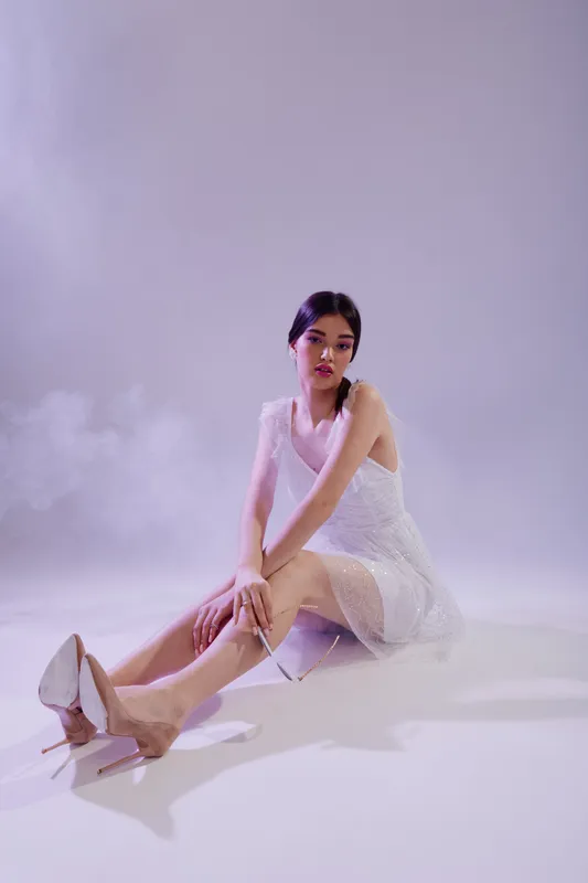 Elegant woman in a white dress and high heels sitting on the floor with a soft purple background. Concept for beauty, fashion, and modern style portrait.