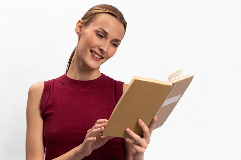 Smiling woman in red sleeveless top reading a book with interest on white background. Concept of education, knowledge, learning, and happiness in a bright studio setting.