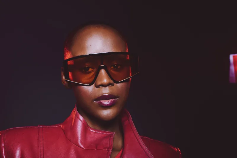Portrait of confident black woman wearing oversized sunglasses and red leather jacket against dark background. Concept of fashion, style, empowerment, and modern urban look.