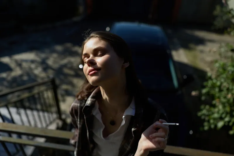 Woman outdoor lifestyle, relaxed and contemplative, sunlit moment on stairs, smoking cigarette, casual jacket, soft breeze, natural tones, moody shadows