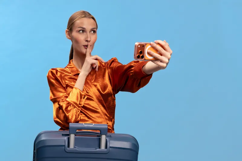 Young woman in orange blouse takes a selfie with a suitcase, making a shushing gesture. Blue background, travel concept and secret vacation idea with casual fashion style.