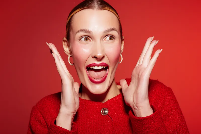 Excited woman in red sweater with open mouth and raised hands expresses surprise and joy on red background. Concept of happiness, emotion, celebration, and positive reaction.