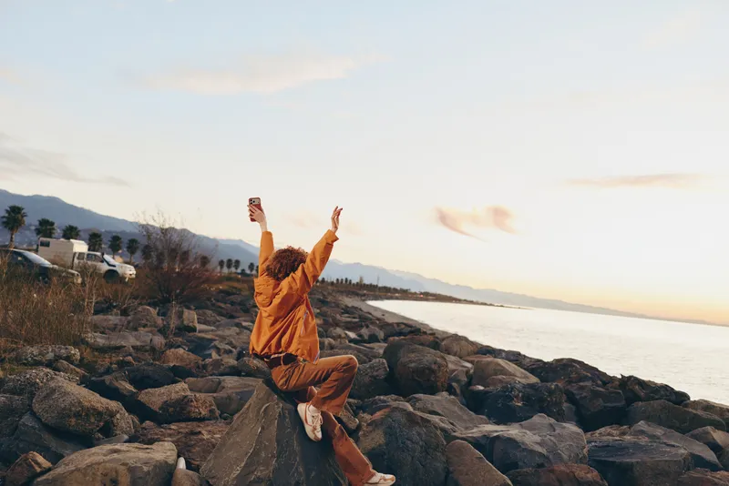 Woman in yoga pose on rocky coast at sunset, outdoors for balance and stretching, seated on stones, calm sea in background, lifestyle wellness concept, warm natural light
